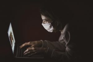 woman in dark with medical mask and gown looking at laptop