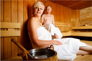 A middle aged woman and man with white towels sit in a wooden sauna.