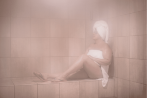 woman sitting in a steam room with a towel on her hair and around her body.